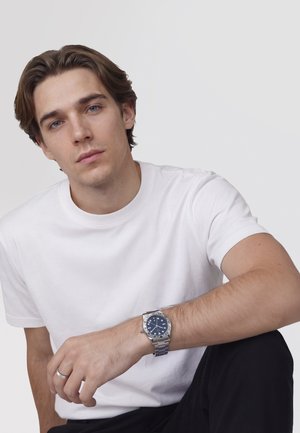 Young man with light brown hair wearing a white t-shirt and silver wristwatch, sitting with one arm resting on his knee against a plain background.