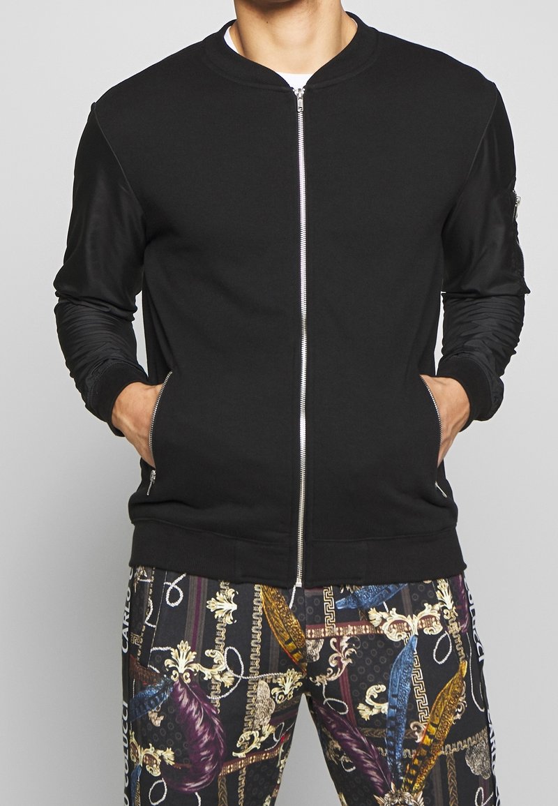 Chaqueta bomber negra con cremallera, puños y cintura acanalados, confeccionada en material suave y con bolsillos laterales, combinada con pantalones estampados.