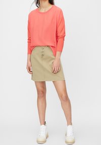 Korálový top s dlouhým rukávem a širokými rukávy v kombinaci s khaki sukní s knoflíky vpředu. Modelka má na sobě bílé vysoké tenisky.