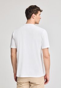 T-shirt bianca in cotone con maniche corte, collo rotondo e una texture liscia. Mostra una vista posteriore semplice su uno sfondo neutro.