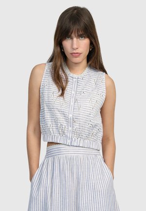 Femme aux longs cheveux bruns portant un haut court sans manches blanc et bleu à rayures avec des boutons et des détails découpés, assorti à une jupe taille haute.