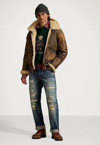 Brown shearling-lined jacket, green graphic sweater, plaid shirt, distressed blue jeans, brown boots with red laces, grey beanie.