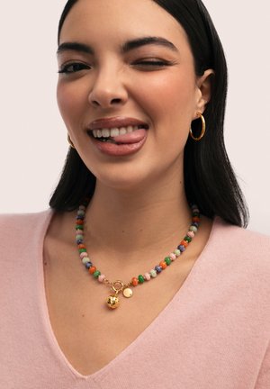 Mujer con cabello largo y oscuro, lleva pendientes de aro dorados y un collar multicolor de cuentas con un charm dorado, guiña un ojo y saca la lengua.