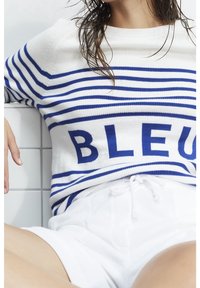Pull en tricot blanc avec des rayures horizontales bleues et le mot "BLEU" en lettres majuscules sur la poitrine. Associé à un short blanc.