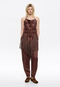 Brown satijnen jumpsuit met bloemvormige borduring, franjes aan de onderkant, zijzakken en taps toelopende pijpen, gecombineerd met bruine suède instapschoenen.