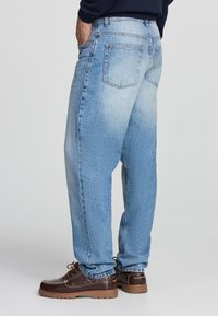 Jeans in denim azzurro chiaro con vestibilità rilassata, lavaggio sbiadito, design a cinque tasche e cuciture a contrasto, abbinati a scarpe marroni.