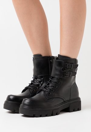 Bottes de combat en cuir noir avec semelles épaisses, lacets à l'avant, fermetures éclair latérales et doubles sangles à boucles, portées sur des jambes nues sur fond blanc.