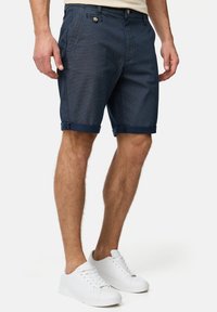 Pantalones cortos de algodón azul marino con un patrón texturizado, puños enrollados, bolsillos delanteros y un cierre de botón único. Llevados con zapatillas blancas.