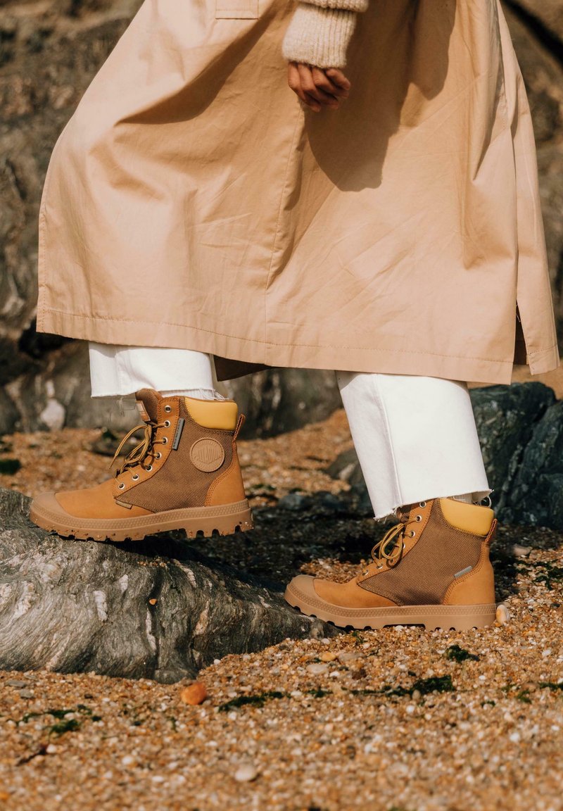 Bottes de randonnée marron avec un tissu texturé et du cuir, accents jaunes et semelles robustes. Portées avec un pantalon blanc et un manteau beige.