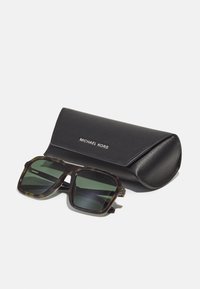 Óculos de sol em tartaruga com lentes verde-escuras, armação quadrada e detalhes em dourado, ao lado de uma capa de couro preto com a inscrição "MICHAEL KORS".