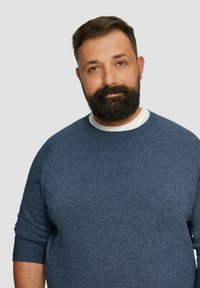 Blauer Strickpullover mit rundem Halsauschnitt und langen Ärmeln, der ein strukturiertes Muster aufweist. Über einem weißen Unterhemd getragen.