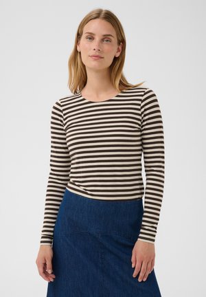 Mujer con cabello castaño claro que lleva una camisa de manga larga a rayas marrón y blanca y una falda vaquera azul, de pie frente a un fondo liso.
