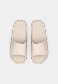 Μπεζ σανδάλια με λουρί, κατασκευασμένα από ομαλό πλαστικό με φαρδύ λουρί και υφασμένο πέλμα, με διακριτικό λογότυπο της μάρκας στην πλευρά.