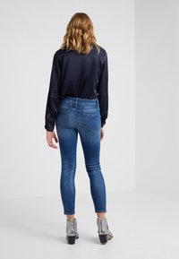 Chemise bleu foncé à manches longues avec des coutures claires, associée à un jean ajusté bleu clair et des bottes argentées à la cheville, vue de dos.