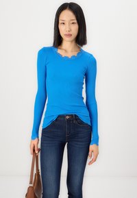Langsleeve ribbed top in helderblauw, met een geschulpte kantnek en manchetten. Gecombineerd met donkere blauwe skinny jeans.