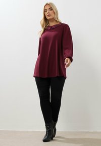 Kvinna som bär en långärmad vinröd tunika med en rutigt krage, svarta smala byxor och svarta klackade ankelstövlar, stående mot en heltäckande vägg.