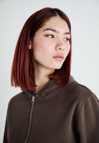 Hnedá mikina na zips s kapucňou. Materiál vyzerá mäkko a štruktúrovane, s koncovkou zipsu v striebornej farbe. Model má vlasy po pleciach v červeno-hnedej farbe.