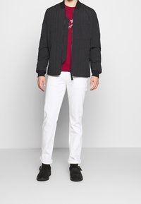 Zwarte gewatteerde bomberjack, rode sweatshirt eronder, witte broek en zwarte instapschoenen. Het jack heeft een frontale rits en ribgebreide manchetten.