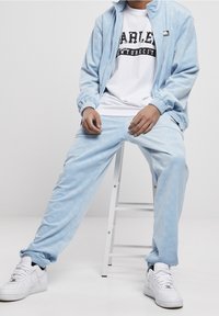 Světle modrá tepláková souprava z měkké, strukturované tkaniny; bunda na zip s vysokým límcem, doplněná o kalhoty ve stejném stylu. Pod tím bílý tričko s potiskem.