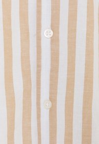 Detailní záběr béžové a bílé látky s vertikálními pruhy, na které jsou uprostřed přišity dva bílé knoflíky, ukazující texturu a detaily šití.