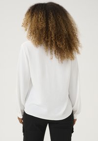 Chemise blanche à manches longues, en tissu léger, avec une coupe décontractée, un ourlet arrondi et des poignets boutonnés. Design simple et minimaliste.