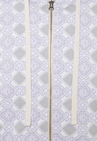 Vêtement zippé avec cordons en tissu blanc sur un motif géométrique pâle de diamants et de formes complexes en gris clair et lavande.