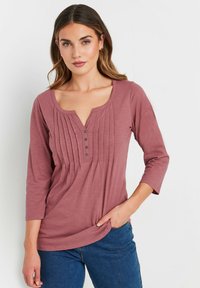 Top lungo color malva a maniche lunghe con scollatura a barchetta, pieghe a pinces sulla parte anteriore e bottoni. Tessuto morbido con vestibilità comoda.