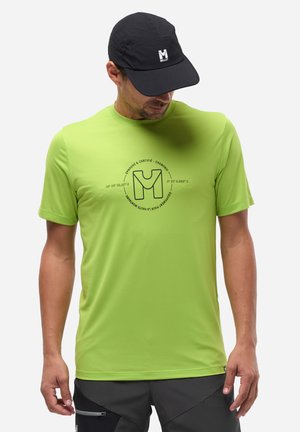 Homme portant une chemise à manches courtes vert vif avec un logo noir et une casquette noire, debout devant un fond clair uni.