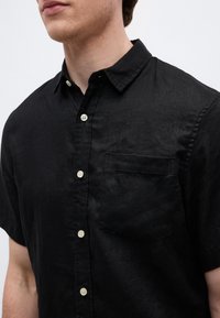 Άνδρας που φοράει μαύρο πουκάμισο με κοντά μανίκια, γιακά και τσέπη στο αριστερό στήθος.