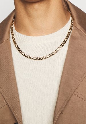 Collier chaîne en or avec des maillons allongés porté sur un pull en maille blanc et sous le col d'un blazer beige au cou d'une personne.