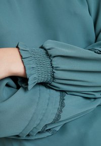 Blouse turquoise avec manches froncées ornées de volants, délicate bordure en dentelle le long de l'ourlet, et une texture de tissu lisse et léger.