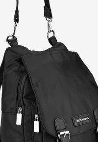 Mochila negra de nailon con solapa plegable, bolsillos con cremallera y acentos de herrajes metálicos. Textura suave con una etiqueta de marca.