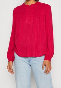 Röd långärmad blus med rund halsringning, knappslå, rynkade axelsömmar och mjukt, texturerat tyg. Matchas med ljusblå jeans.