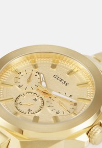 Gouden metalen horloge met een gestructureerde wijzerplaat, drie subwijzerplaten en opvallende uurmarkeringen. Het merk "GUESS" is op de wijzerplaat weergegeven.