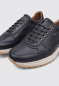 Un par de zapatillas de cuero azul marino con cordones negros, medias suelas blancas y suelas exteriores de goma marrón claro sobre un fondo plano.
