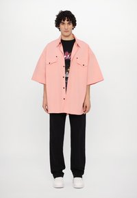 Camisa rosa claro de gran tamaño con mangas cortas, bolsillos delanteros y cierre de botones, llevada sobre una camiseta gráfica negra y combinada con pantalones negros.