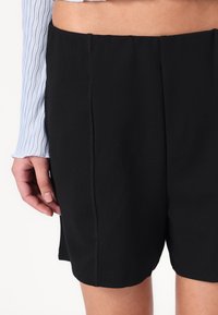 Sorte shorts med en glat tekstur, med lige snit og synlige sidesømme; bæres med en lys blå, struktureret langærmet top.