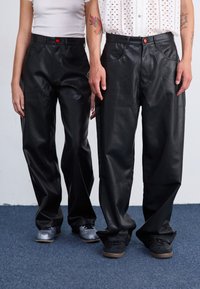 Zwarte leren broek met een losse pasvorm, voorzien van voorzakken en een rode knopaccent. Gedragen met grijze en zwarte schoenen.