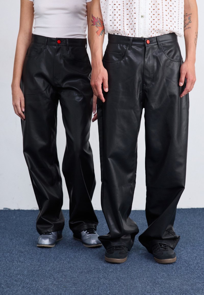 Zwarte leren broek met een losse pasvorm, voorzien van voorzakken en een rode knopaccent. Gedragen met grijze en zwarte schoenen.