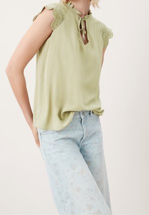 Kvinde iført lys grøn ærmeløs bluse med blonde skuldre og lyseblå jeans med blomsterprint, hånd hvilende på hoften.