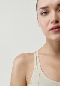 Beige top s tenkými ramínky, s hladkou texturou a minimálním šitím. Detailní záběr na výstřih a oblast ramen.