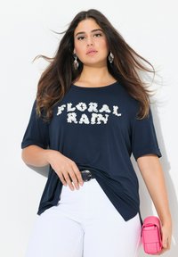 Námořnická modrá tričko s krátkým rukávem a bílým květinovým nápisem "FLORAL RAIN"; spárováno s bílými džínami a růžovou kabelkou.