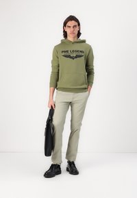 Oliwkowa bluza z kapturem z czarnym tekstem i grafiką skrzydeł, jasnoszare spodnie, czarne buty oraz czarna torba. Czysty, minimalistyczny design.