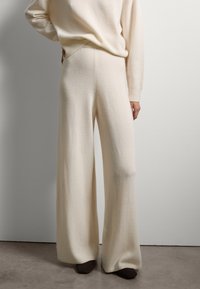 Wijdvallende broek van zacht wit gebreid materiaal, hoog in de taille met een gladde textuur, voorzien van een geribbelde tailleband en boorden. Vergezeld van donkere schoenen.