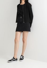 Černá texturovaná bomber bunda přes černý top, doplněná o černou džínovou mini sukni. Celý outfit zakončují černé tenisky s bílými akcenty.