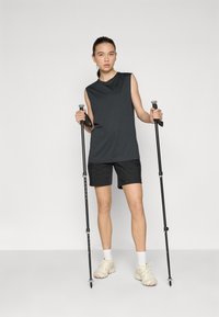 Mørk grå ærmeløs topp og svarte shorts, holder svarte vandrestaver, står på en lys grå bakgrunn. Aktivt tøy-design, myk tekstur.