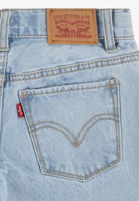 Jeans in denim blu chiaro con una leggera texture, caratterizzati da un'etichetta di marca in pelle marrone e una iconica linguetta rossa sulla tasca posteriore.
