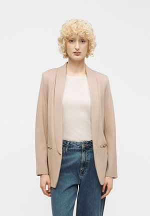 Personne aux cheveux blonds bouclés courts portant un blazer beige, un haut blanc et un jean bleu, debout devant un fond blanc uni.