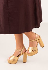 Sandalias de tacón alto doradas con tiras trenzadas y plataforma. Correas cruzadas y detalle de hebilla en el tobillo, con un acabado suave.