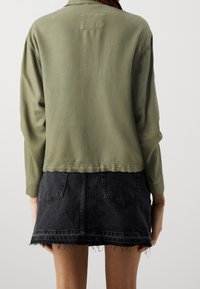 Camicia verde oliva a maniche lunghe con taglio dritto, abbinata a una mini gonna di denim nera con orlo sfrangiato e tasche posteriori.